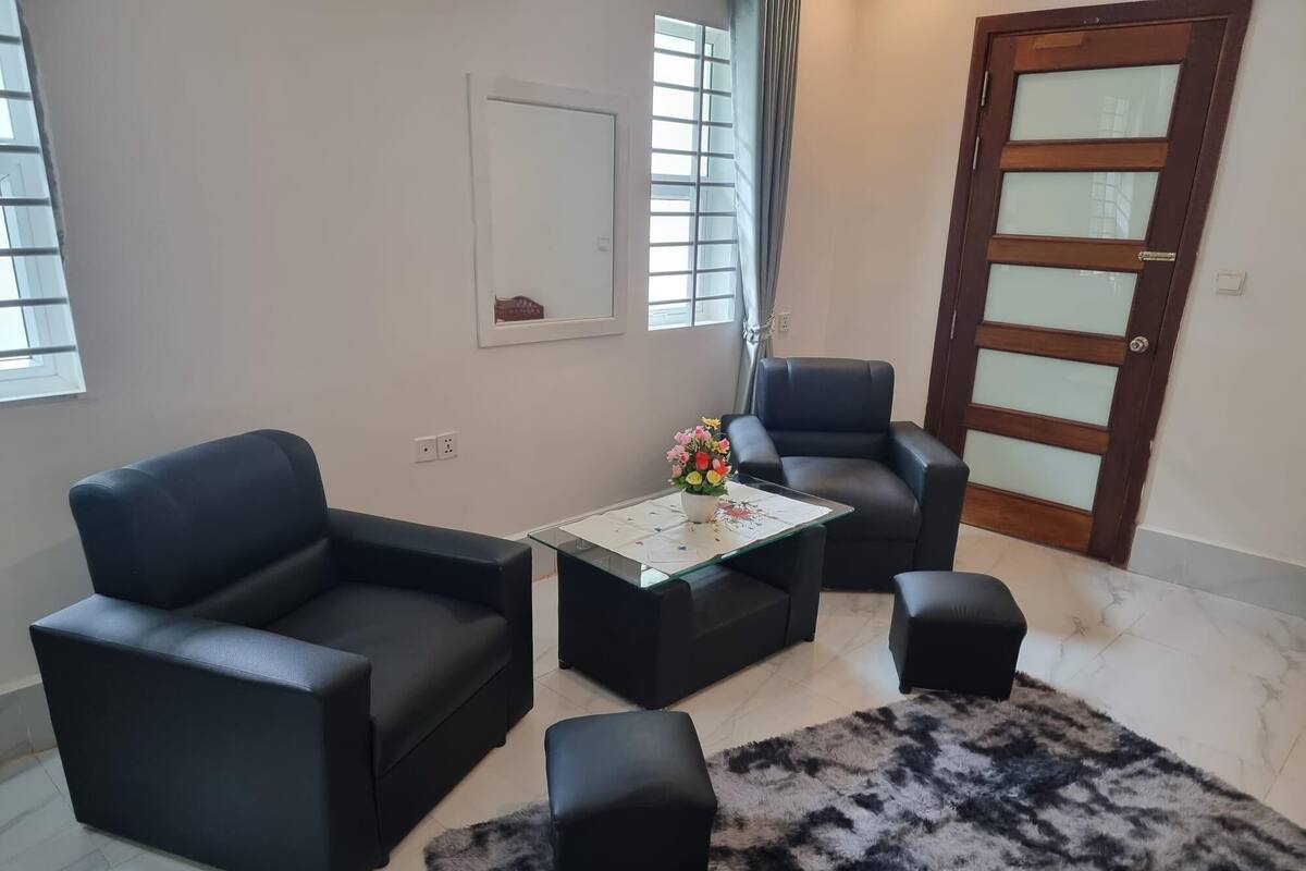 Een woonkamer met een zwarte leren bank, een bijpassende fauteuil, een glazen salontafel met een bloemstuk en een zwart pluchen vloerkleed.
