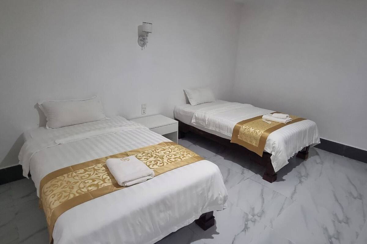 Twee enkele bedden met witte beddengoed en gouden accenten, een wandlamp en een marmeren vloer.