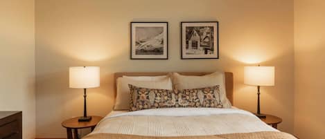 Quarto de hotel com cama, duas luminárias de cabeceira, mesa de cabeceira e duas fotos emolduradas na parede.