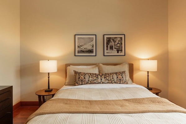 Habitación de hotel con cama, dos lámparas de noche, una mesita de noche y dos cuadros en la pared.