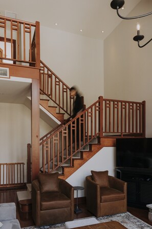 Una escalera con barandillas de madera que conduce a un piso superior, un televisor de pantalla plana y dos sillones de cuero marrón en una sala de estar.