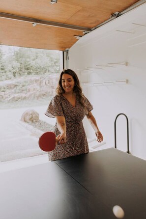 Una persona jugando ping pong en una habitación con un ventanal grande, una mesa de ping pong y una silla.