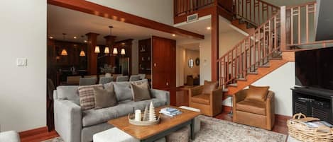 A spacious living area with a grey sofa, brown armchairs, a wooden coffee table, and a staircase with wooden railings.
