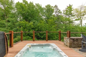 Un jacuzzi en una terraza de madera, con valla de madera y pared de piedra, rodeado de frondosos árboles verdes.