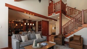 A spacious living room with a grey sofa, brown armchairs, and a wooden coffee table. A staircase with a wooden railing leads to an upper level. - HOT TUB - Ping-Pong - BBQ Terrace - Free Shuttle (Mont-Tremblant)