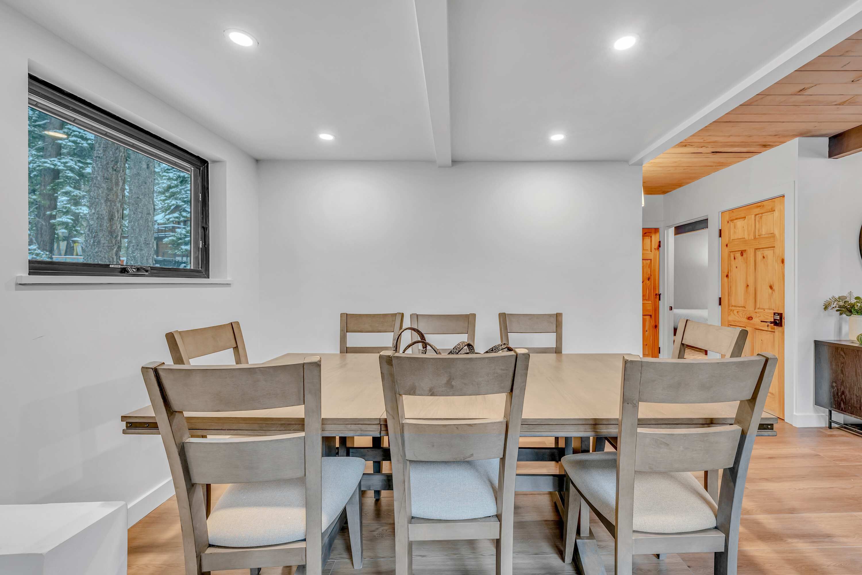 Un coin repas avec une grande table en bois et six chaises, une fenêtre donnant sur les arbres et un plafond en bois.