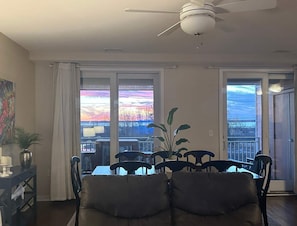 A dining area with a table and chairs, a ceiling fan, and large windows with a view of the sky.