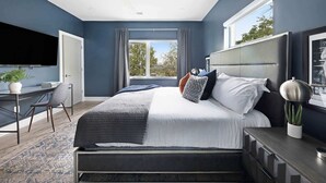 A modern bedroom with a large bed, a desk with a chair, a ceiling fan, and a TV mounted on the wall.
