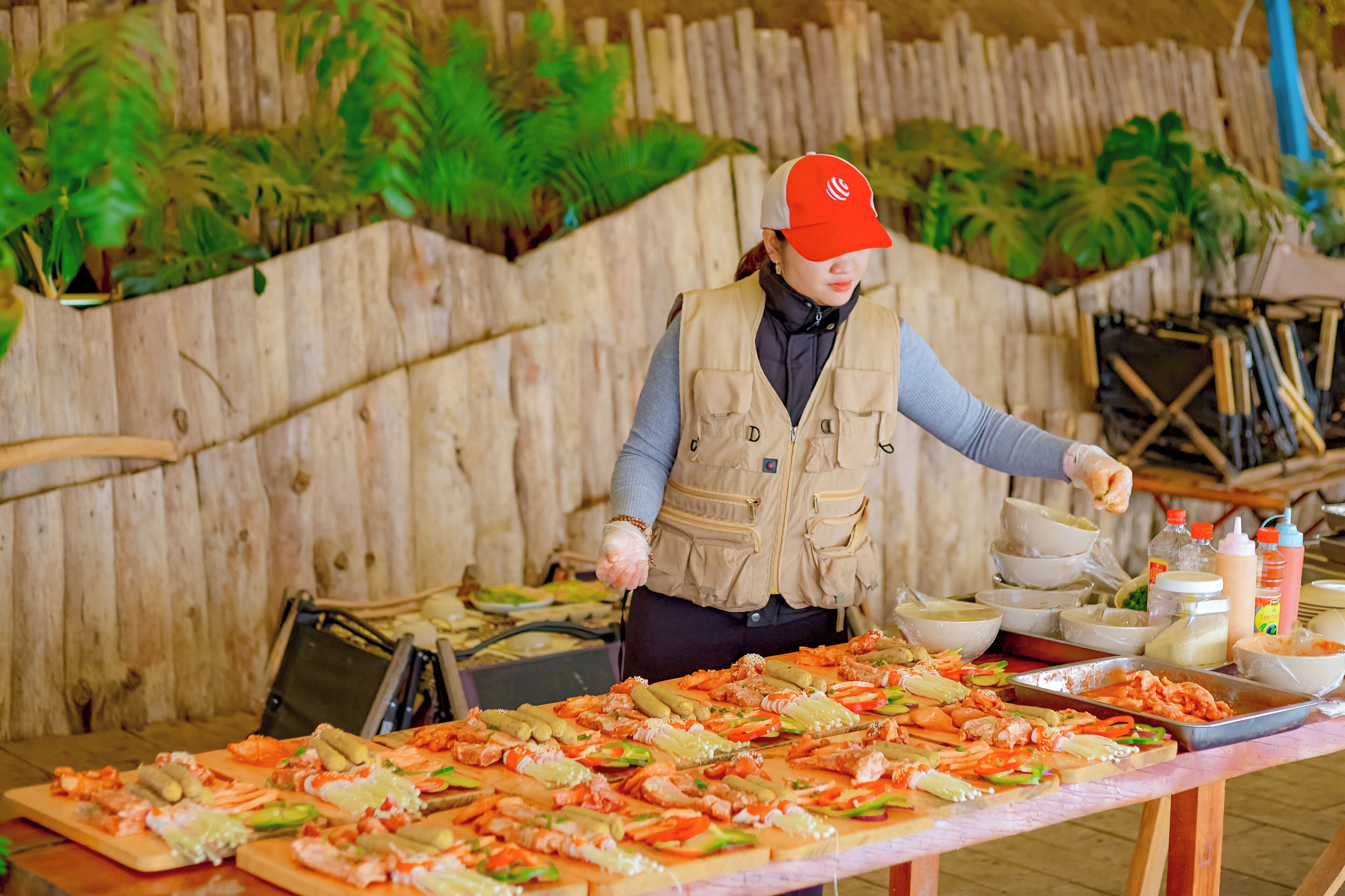 A buffet table with various dishes, a person serving food, and a wooden wall with plants in the background.