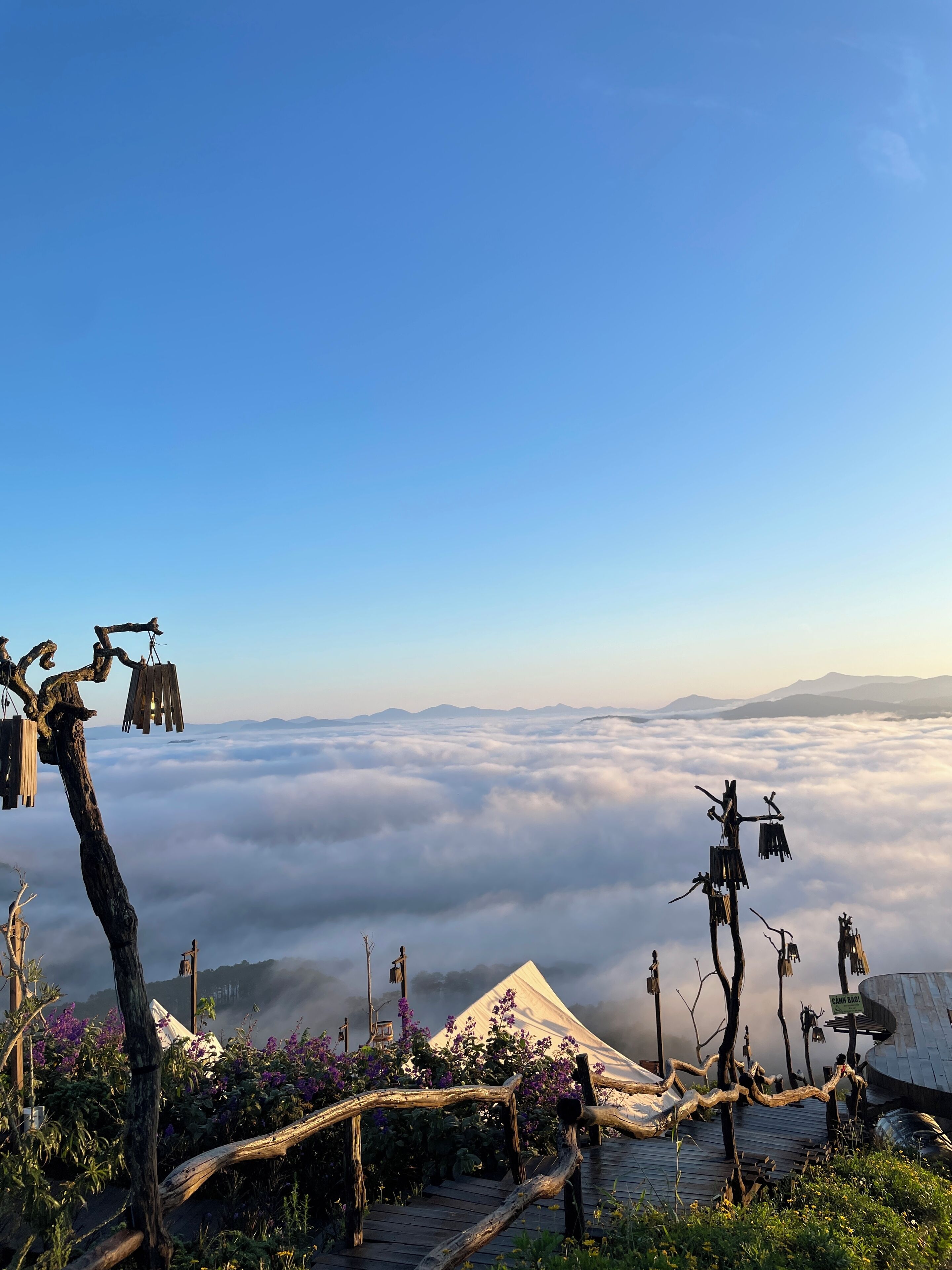 A high-altitude view with a wooden bridge and rustic structures, surrounded by clouds and mountains.