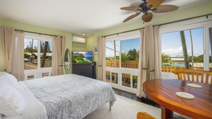Un dormitorio con una cama, una mesa de madera con sillas, un ventilador de techo y ventanas grandes con cortinas.