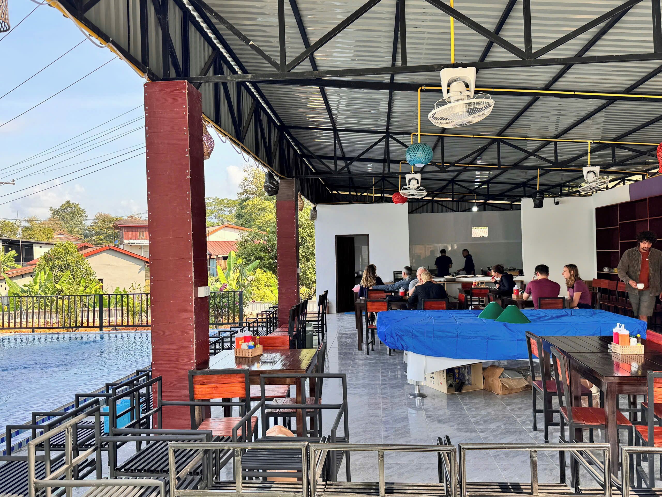 An outdoor restaurant with tables and chairs, a pool in the background, and a covered seating area with a blue tablecloth.