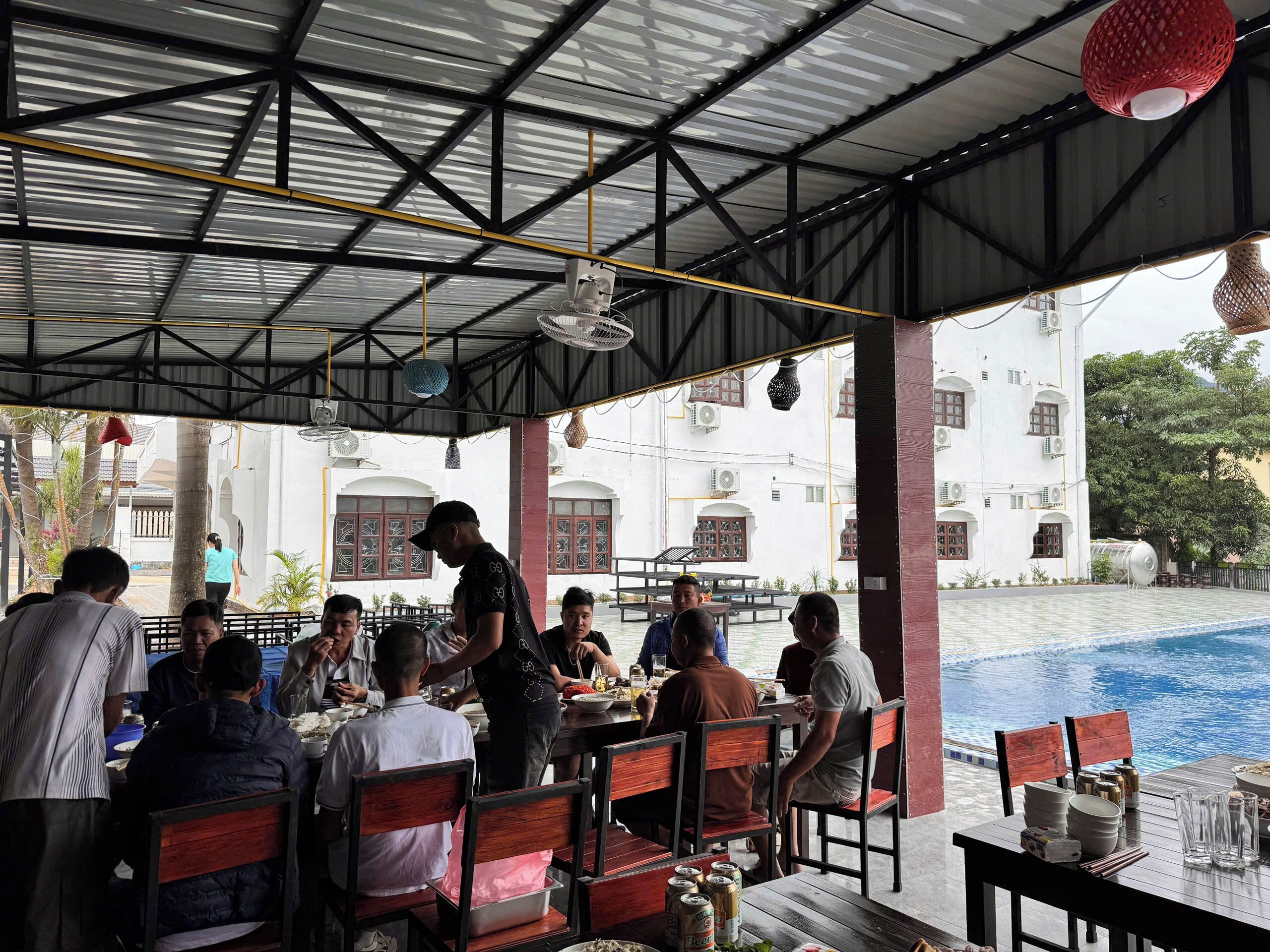 Un restaurant en plein air avec des tables et des chaises, une piscine en arrière-plan et un espace couvert doté d’un toit en métal.