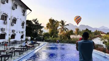 A pool area with a view of mountains, a building with arched windows, and a hot air balloon in the sky.