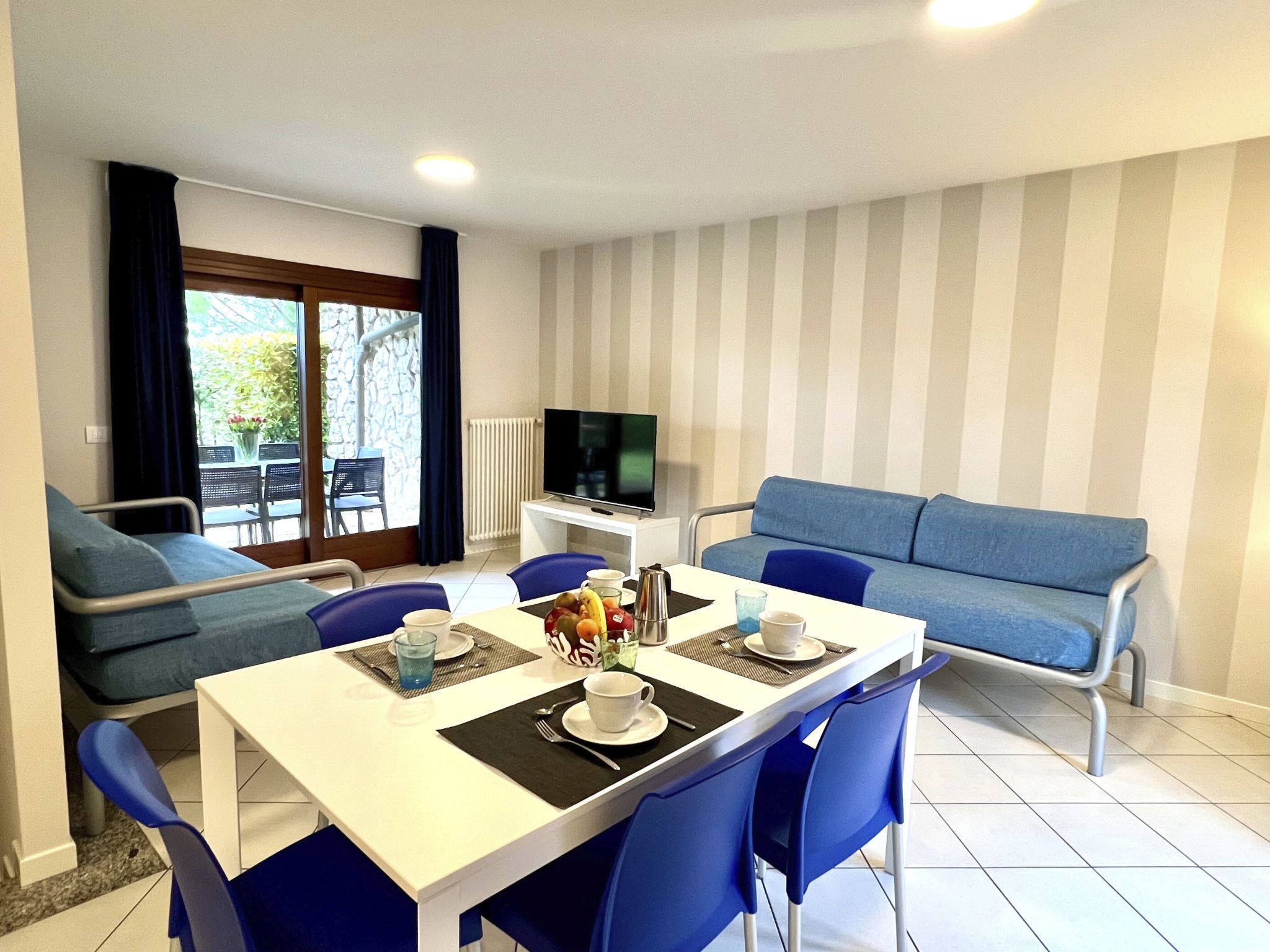 A dining area with a table set for two, a blue sofa, a TV, and a view of the outdoors.