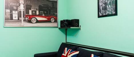 Una habitación con una pared azul aguamarina, un sofá negro con dos cojines con la bandera Union Jack, un anuncio antiguo de una tienda General enmarcado, una fotografía de un paisaje urbano enmarcada, y un altavoz en un estante.