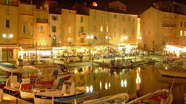 Una marina de noche con barcos amarrados, edificios con ventanas iluminadas y una superficie de agua tranquila que refleja las luces.