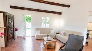Amplia sala de estar con un sofá grande, una mesa de centro y un sillón. Hay una puerta de madera y una ventana con vista a zonas verdes.