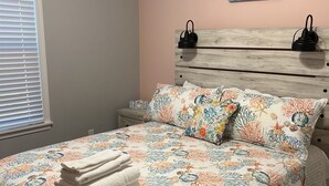 Ein Schlafzimmer mit einem Bett, einem Nachttisch, einem Wandbild und einem Fenster mit Jalousien.