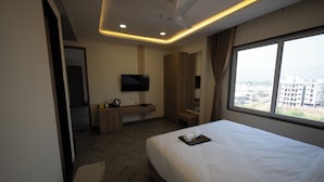 A hotel room with a bed, a desk with a TV, a window with a city view, and a ceiling fan. - Lotus Leaf (Sudhagad)