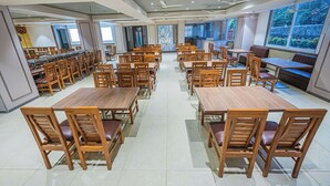 A spacious dining area with multiple wooden tables and chairs, large windows offering views of greenery, and a modern ceiling design. - Lotus Leaf (Sudhagad)