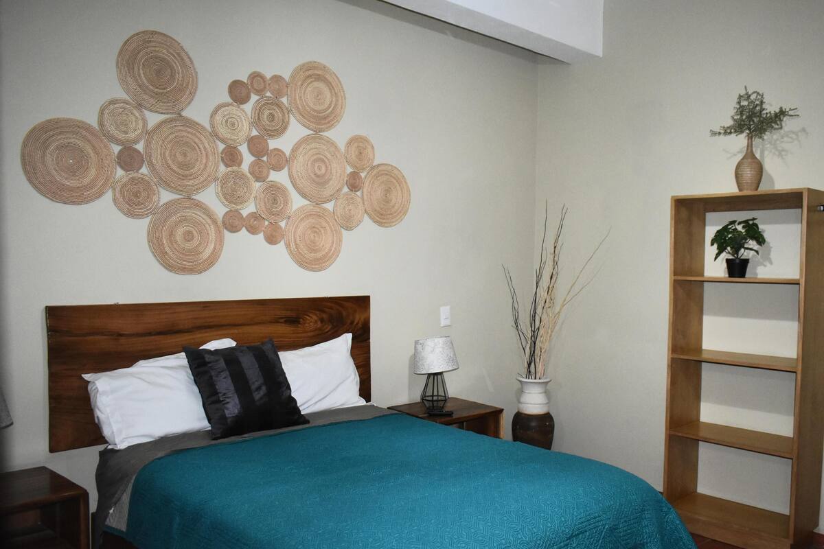 A neatly arranged bedroom with a bed, a bedside table, a wall art piece, a vase with branches, and a wooden shelf with a plant.