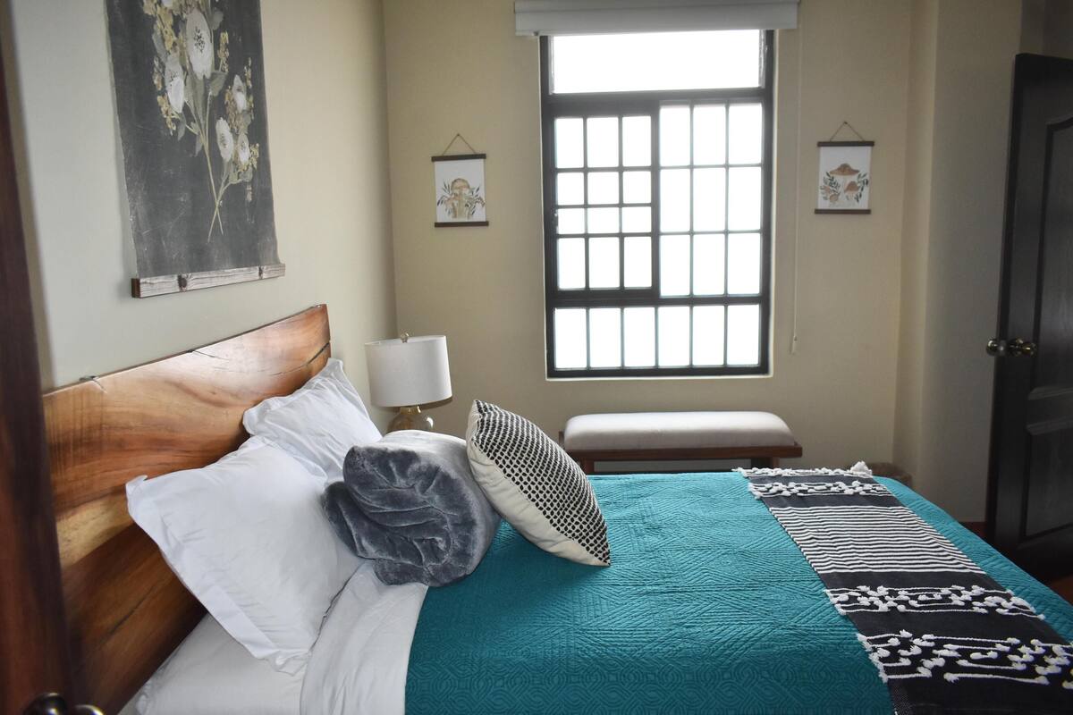A neatly made bed with a teal bedspread and patterned pillows, a bedside lamp, and a window with a view of the outside.