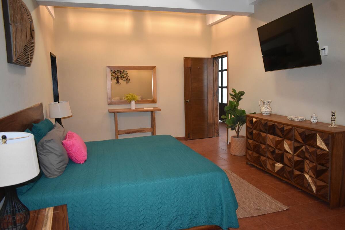 A bedroom with a bed, bedside table, lamp, dresser, TV, and a door leading to another room.