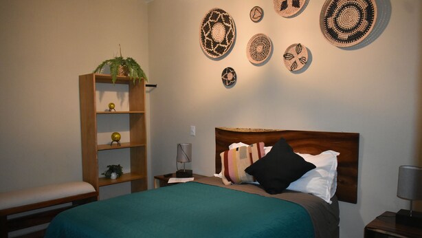 A bedroom with a bed, a nightstand, a lamp, a bench, a shelf with decorative items, and wall art. - Hotel Boutique Casa Tastuan (Tonalá)