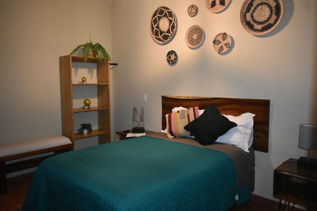 A bedroom with a bed, a nightstand, a lamp, a bench, a shelf with decorative items, and wall art.