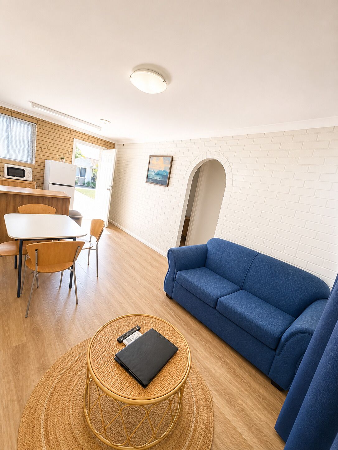A compact living space with a kitchenette, a dining area, a sofa, and a round wicker table with a remote control and a book.
