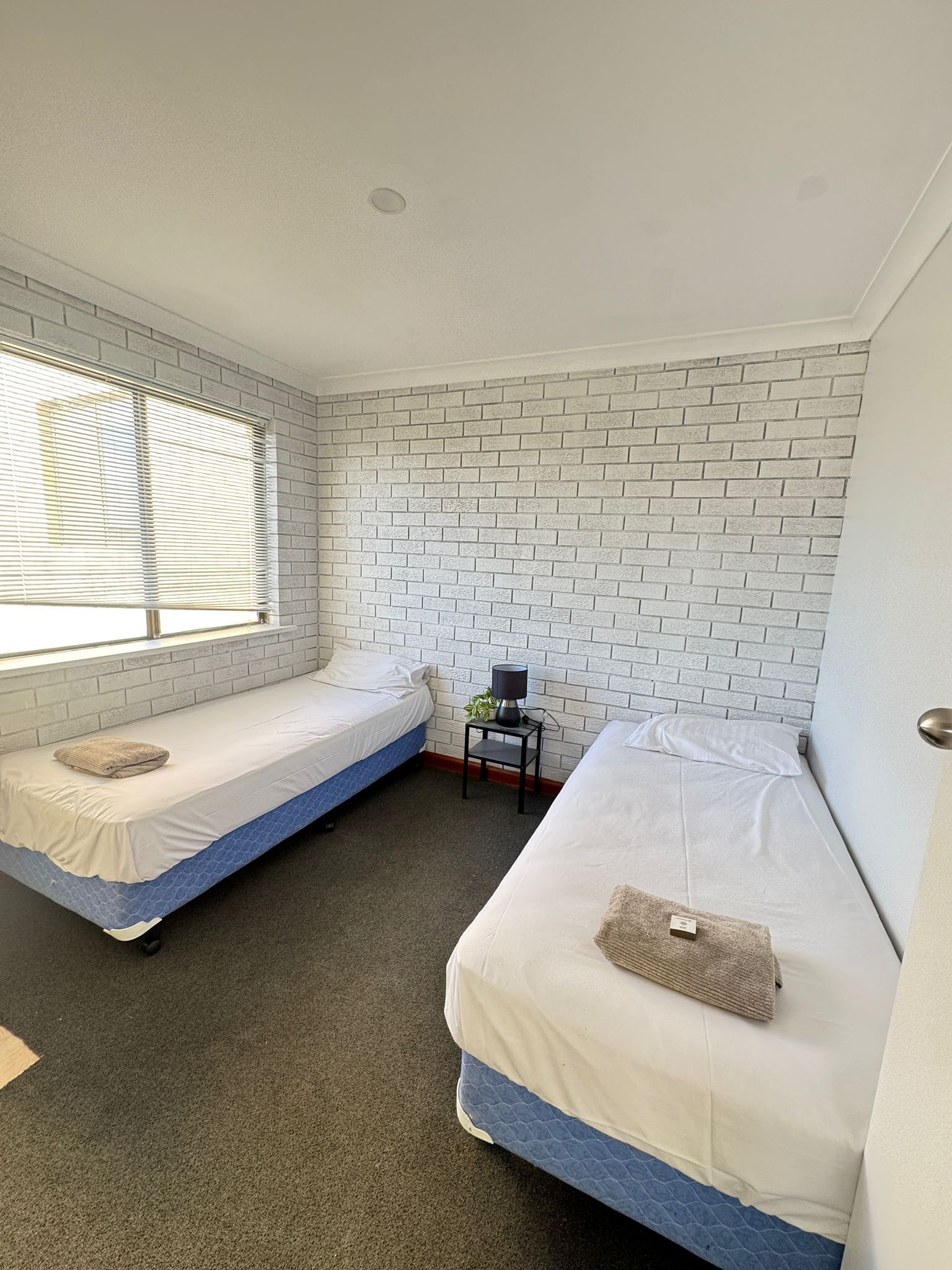A small, clean room with two single beds, a small table with a lamp, and a window with blinds.