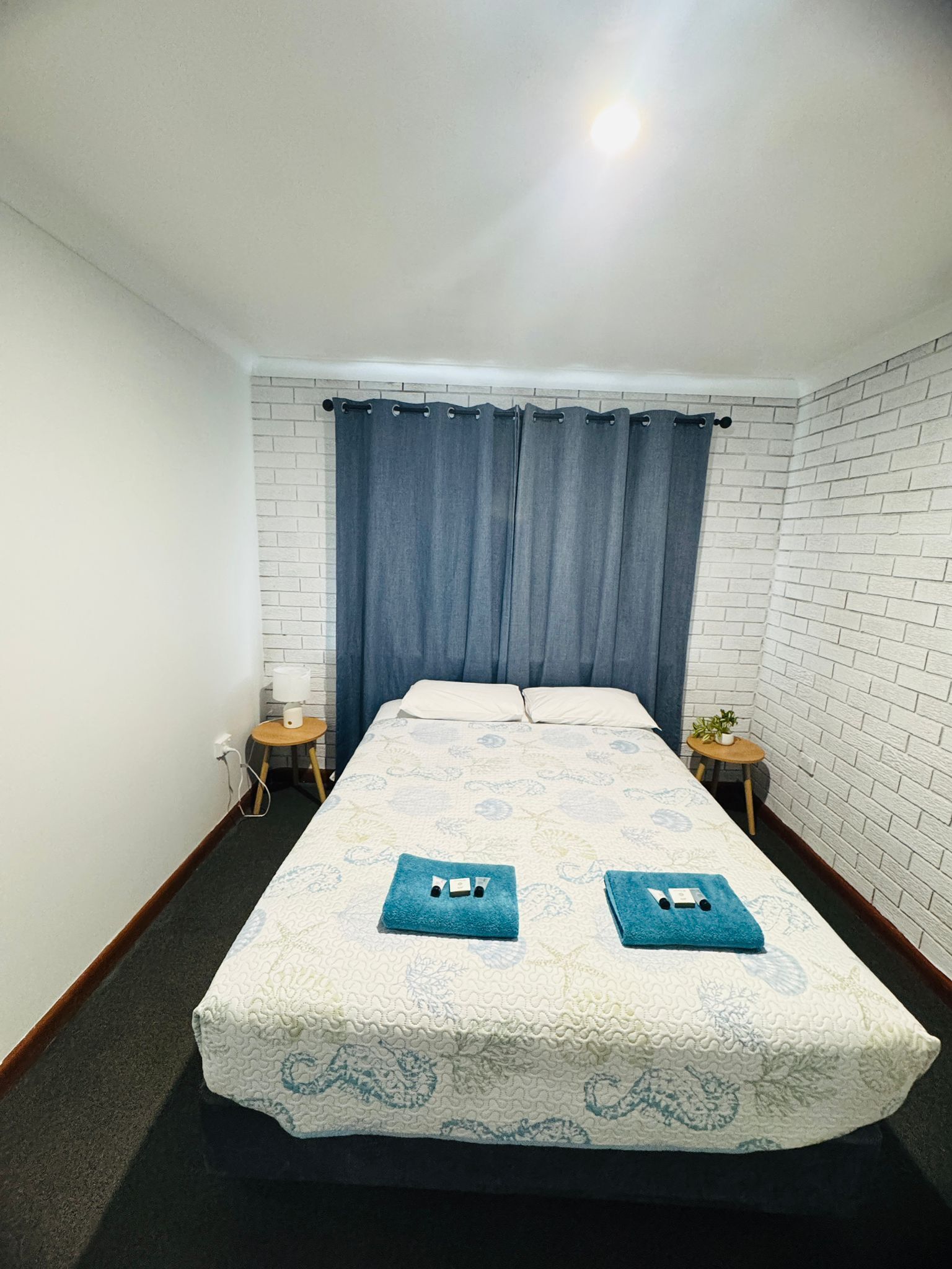 Un dormitorio con una cama, una mesita de noche, una lámpara, una pequeña mesa con una planta y una cortina azul.