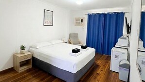 Ett hotellrum med en säng, ett litet sidobord med en växt, en stol, ett skrivbord med en dator och en blå gardin.