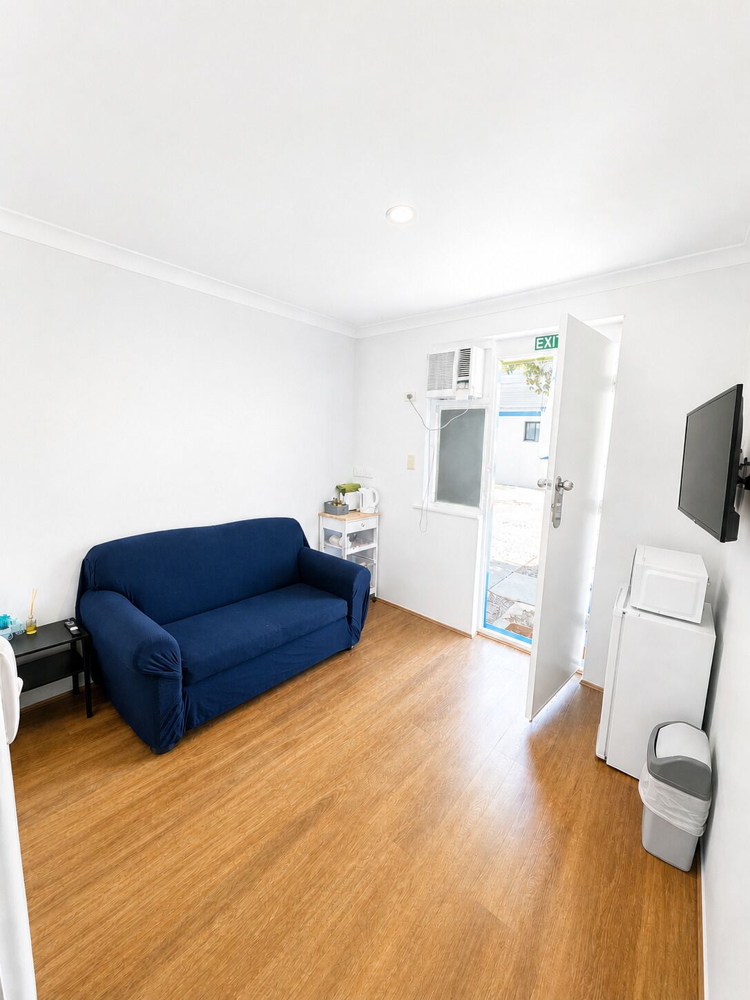 A room with a blue sofa, a small table with items, a white door with an exit sign, a white refrigerator, and a trash bin.