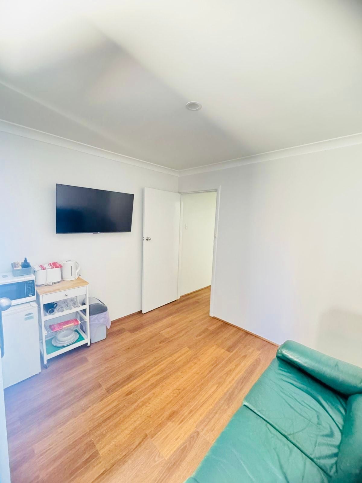 A room with a flat-screen TV mounted on the wall, a white cabinet with kitchen appliances, a green sofa, and a wooden floor.