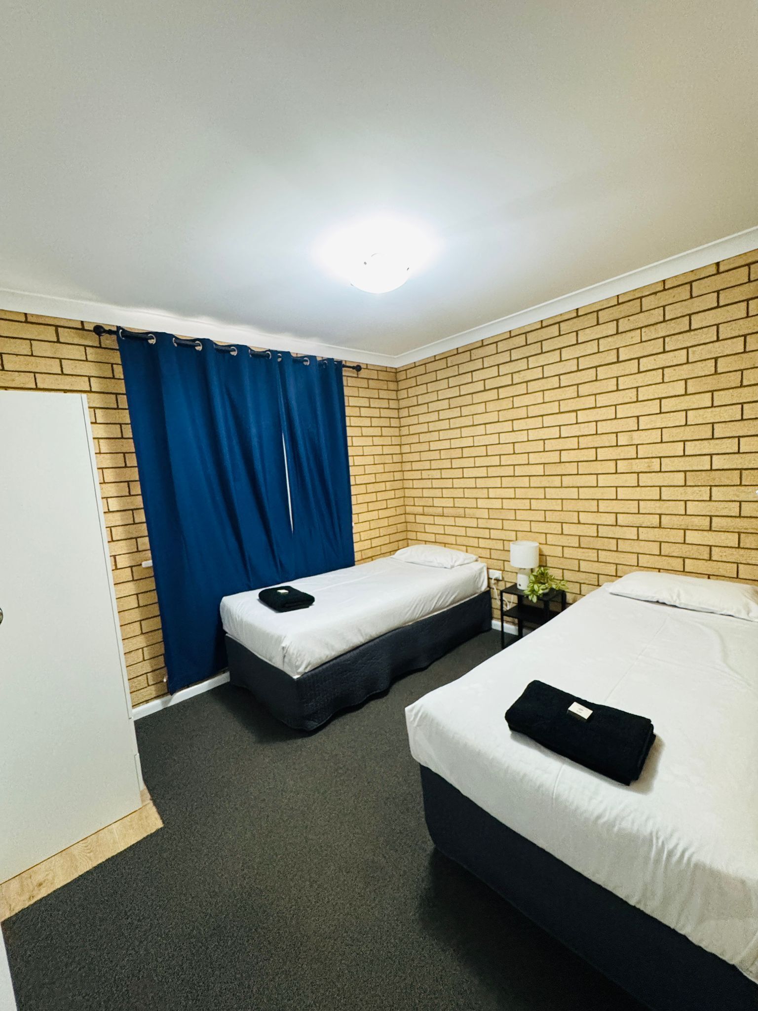 A hotel room with two beds, a brick-patterned wall, a blue curtain, a white door, and a small bedside table with a lamp.