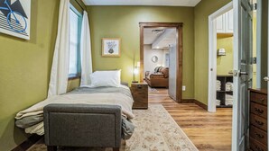 Un dormitorio con cama, mesita de noche y tocador. Hay una ventana con cortinas, un cuadro enmarcado en la pared y una puerta que da a otra habitación.