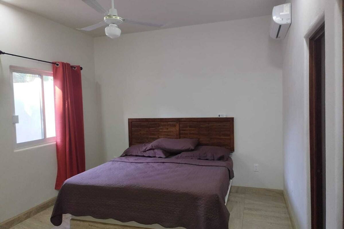 Een slaapkamer met een bed, een plafondventilator, een raam met rode gordijnen en een airconditioning.