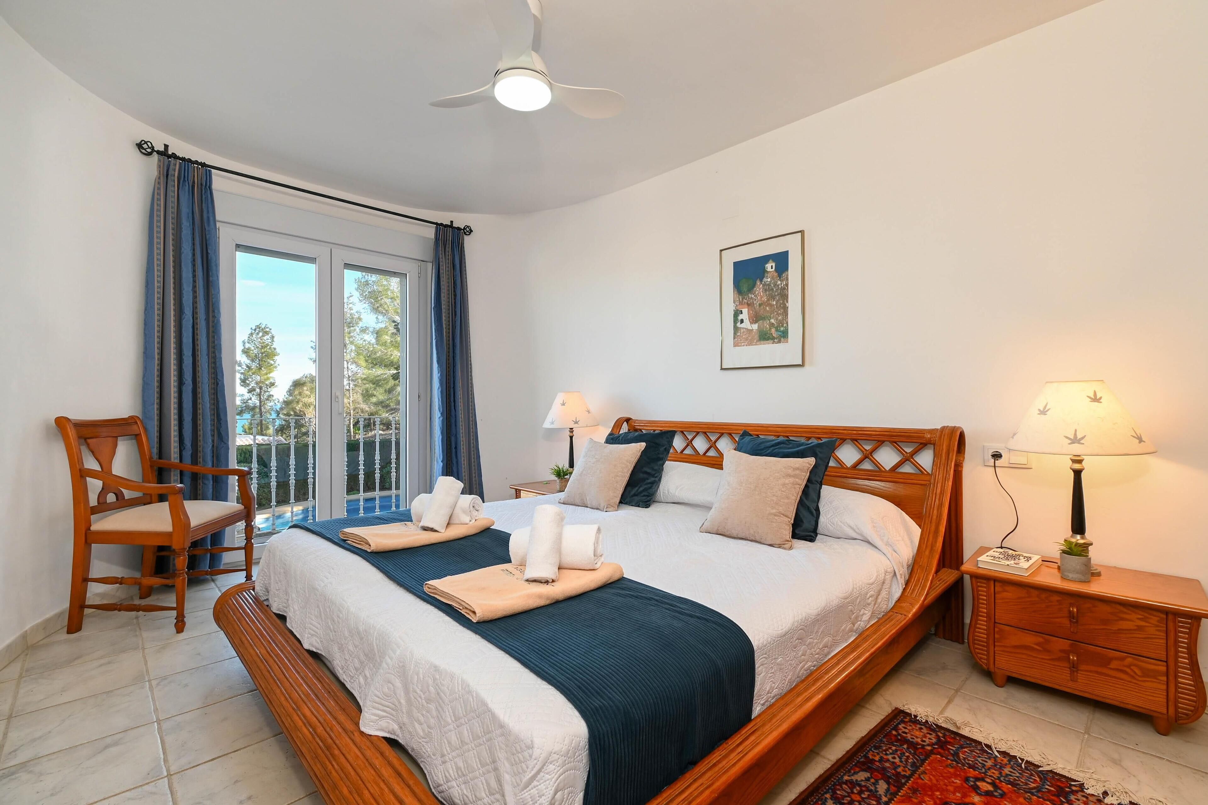 A bedroom with a large bed, a wooden chair, a nightstand, and a view through a window.
