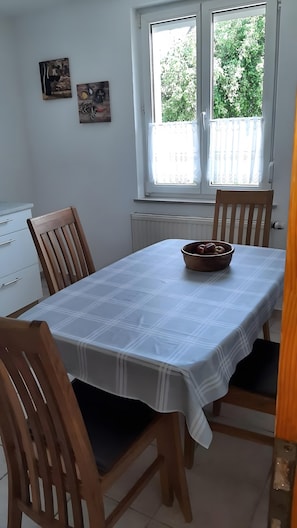 Una mesa de comedor con mantel blanco, sillas de madera y un bol sobre la mesa. Se observan dos ventanas con cortinas, un radiador debajo y cuadros colgados en la pared.