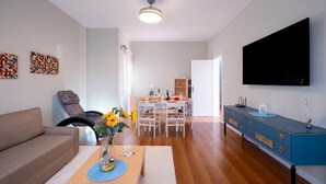 Un salon avec un canapé, un coin repas avec une table dressée pour deux personnes, un meuble bleu et un téléviseur à écran plat fixé au mur.