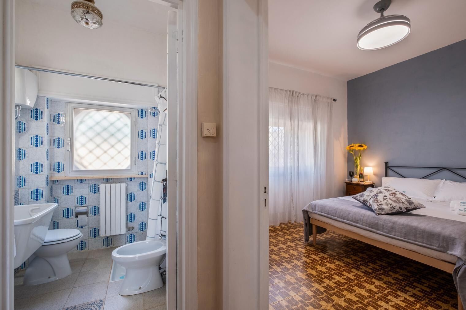 Quarto compacto com cama, mesa de cabeceira com luminária e janela com cortinas finas.
