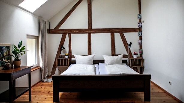 A bedroom with a wooden ceiling, a bed with white bedding, a nightstand, a potted plant, and a window with curtains.