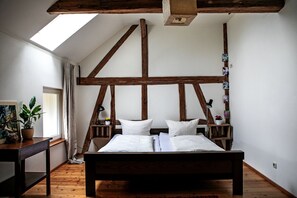 Un dormitorio con techo de madera, una cama con ropa blanca, una mesita de noche, una planta en maceta y una ventana con cortinas.