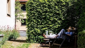 A person relaxing on a chair in a garden with a large hedge and a building in the background.