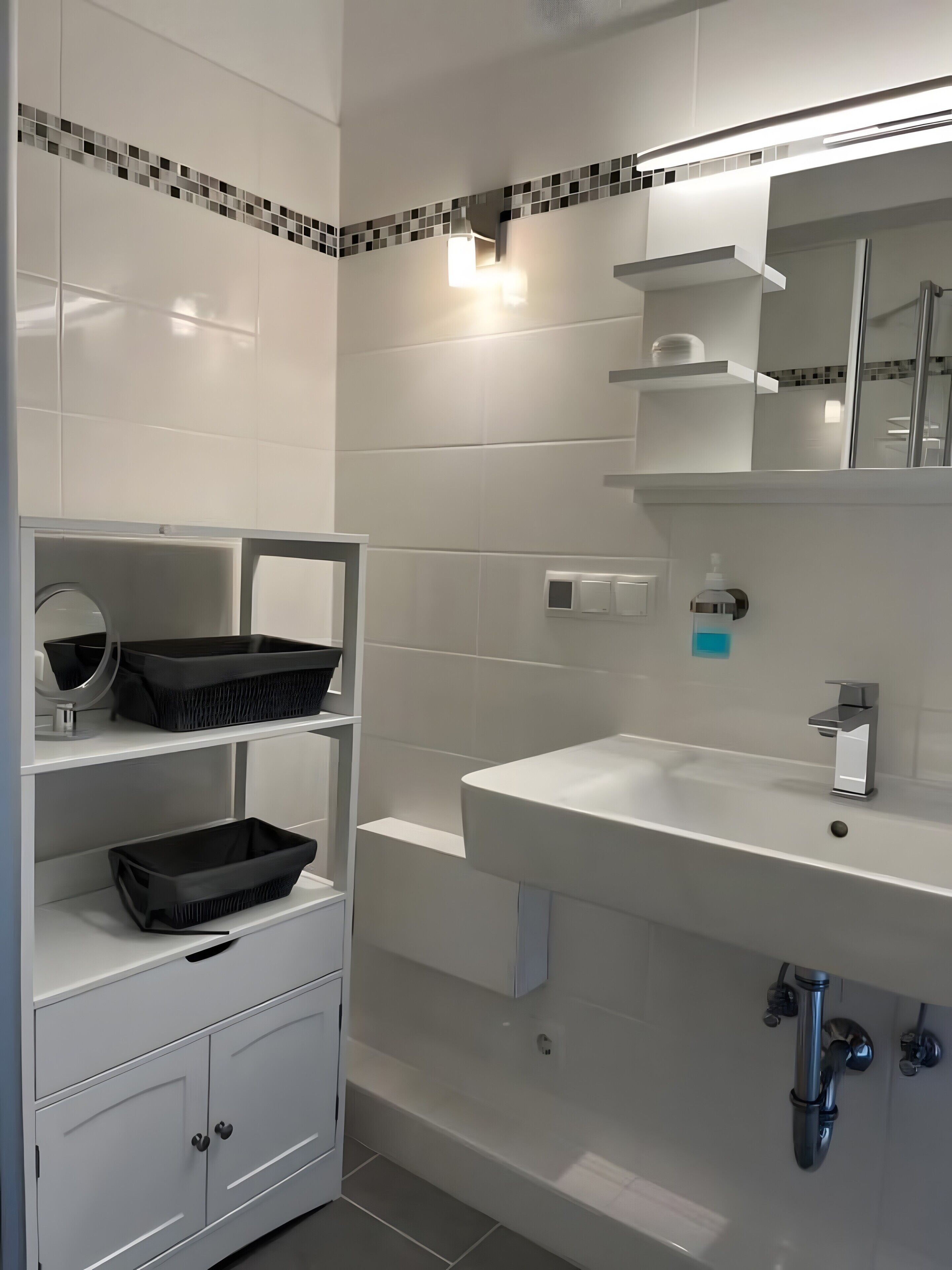 A modern bathroom with white tiles, a white sink, a mirror, and a shelf with black baskets.