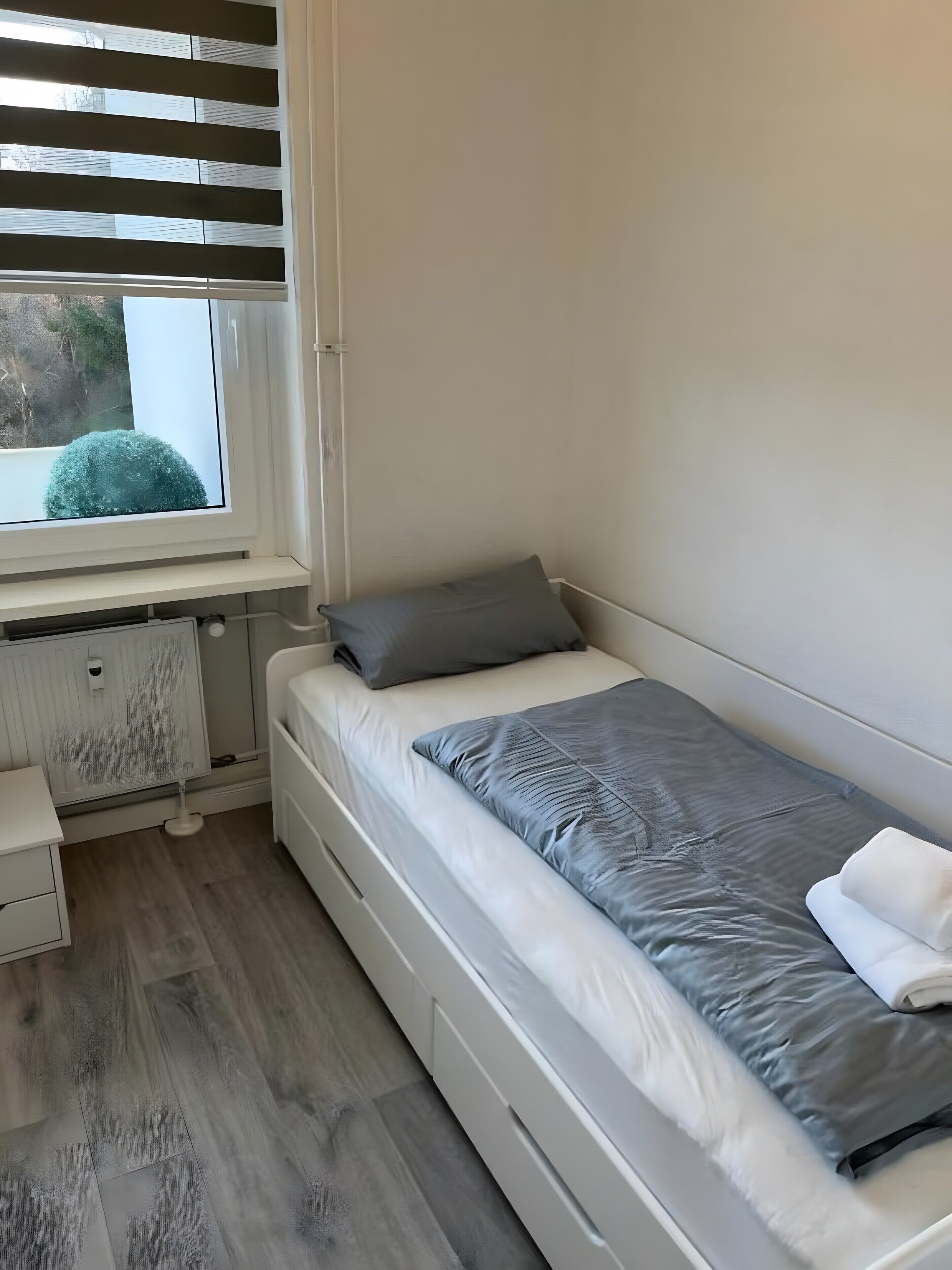 A small, single-bed room with a window, a radiator, a bedside table, and a view of greenery outside.