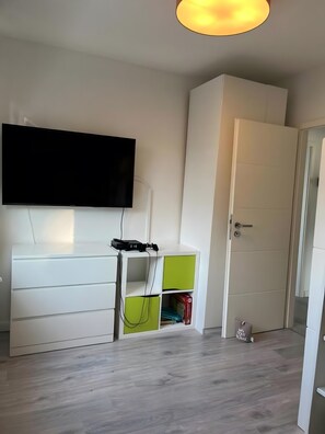 A room with a flat-screen TV mounted on the wall, a white wardrobe, a green storage unit, and a door.