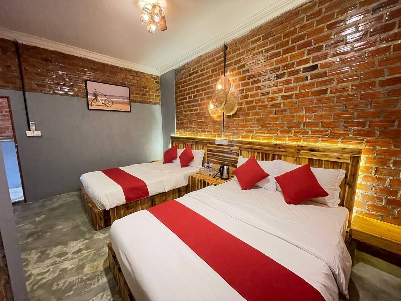 A hotel room with two beds, each with white bedding and red accents, a brick wall, and a ceiling light fixture.
